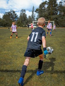 Nuori poika, maalivahti on potkaisemassa jalkapalloa ja muut odottavat sitä kauempaana.